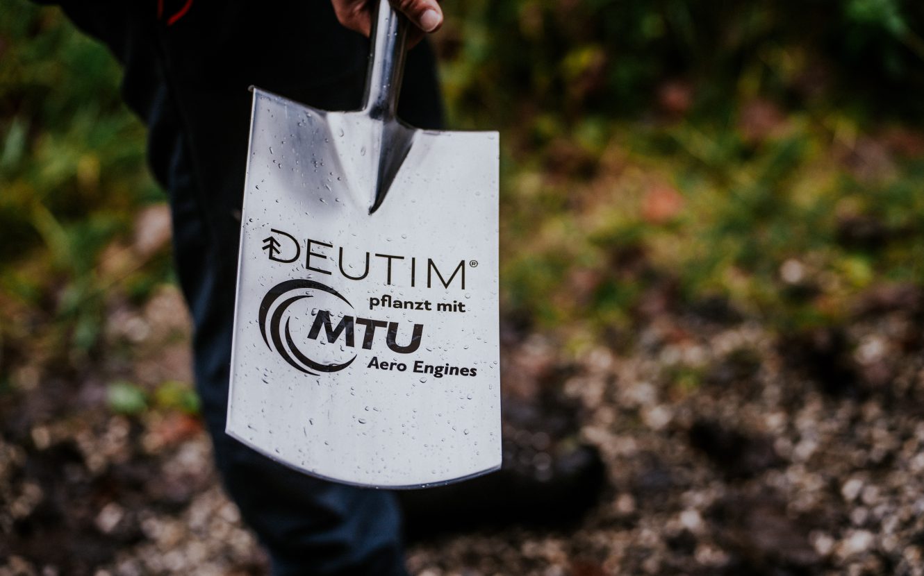 Nahaufnahme. Person hält Edelstahlspaten mit Deutim- und MTU Logo.